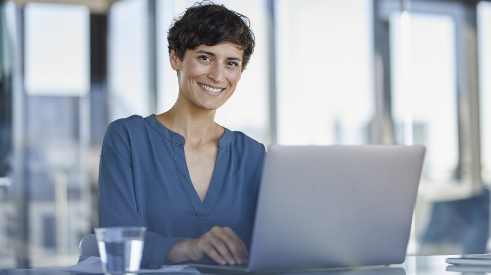Eine Frau im blauen Shirt sitzt mit Laptop im Büro und betreibt Active Sourcing.