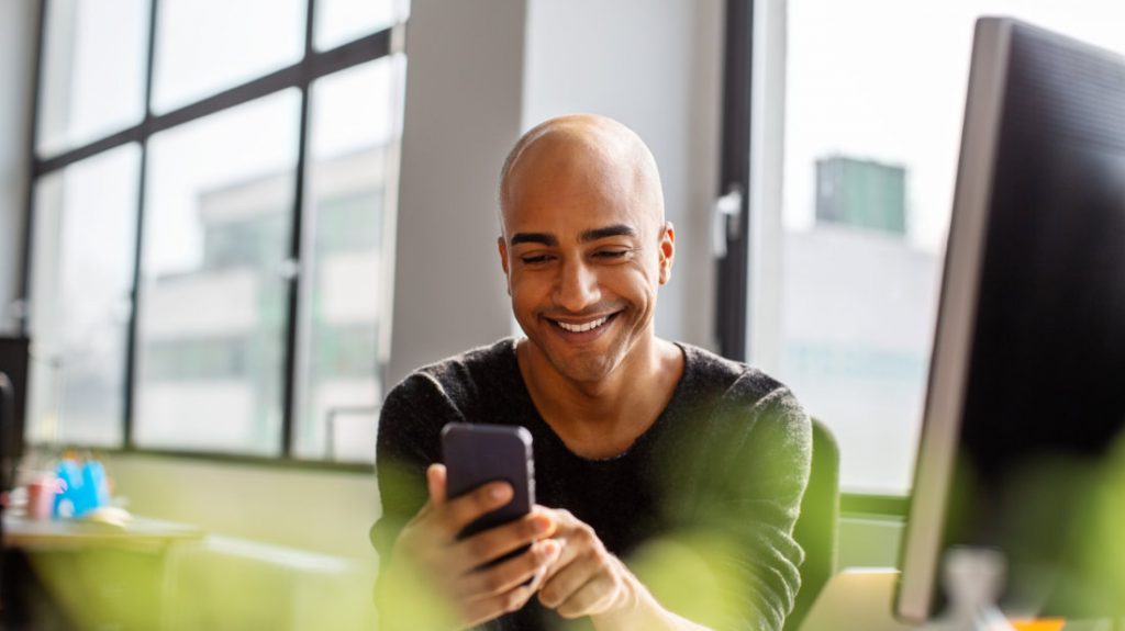 Ein Mann mit seinem Smartphone in der Hand sitzt im Büro vor seinem PC und nutzt Social Media im Recruiting. 